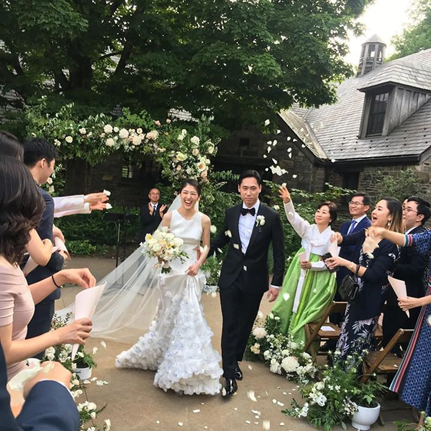 NYC weddinf DJ Bride and groom walk under a flower arch as guests cheer and toss petals at an outdoor wedding with stone buildings and trees