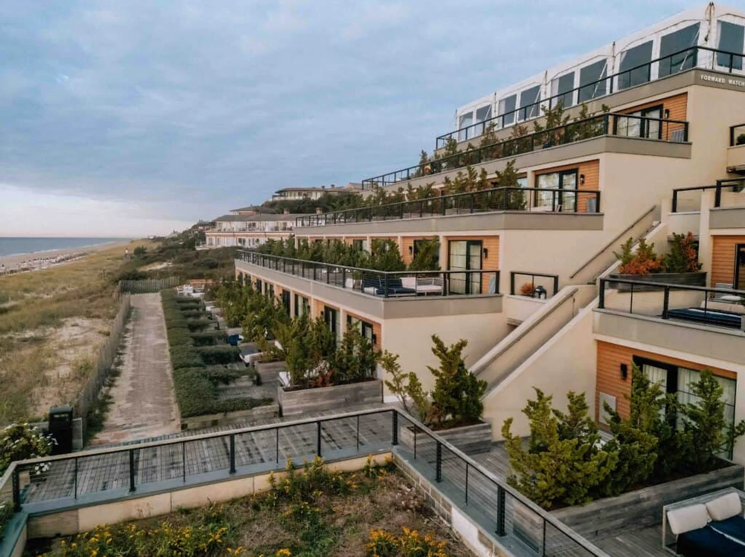 NYC Wedding DJ Bride Modern seaside resort apartments on dunes under cloudy sky, with balconies and beach path; faint text reads FORWARD WATCH.