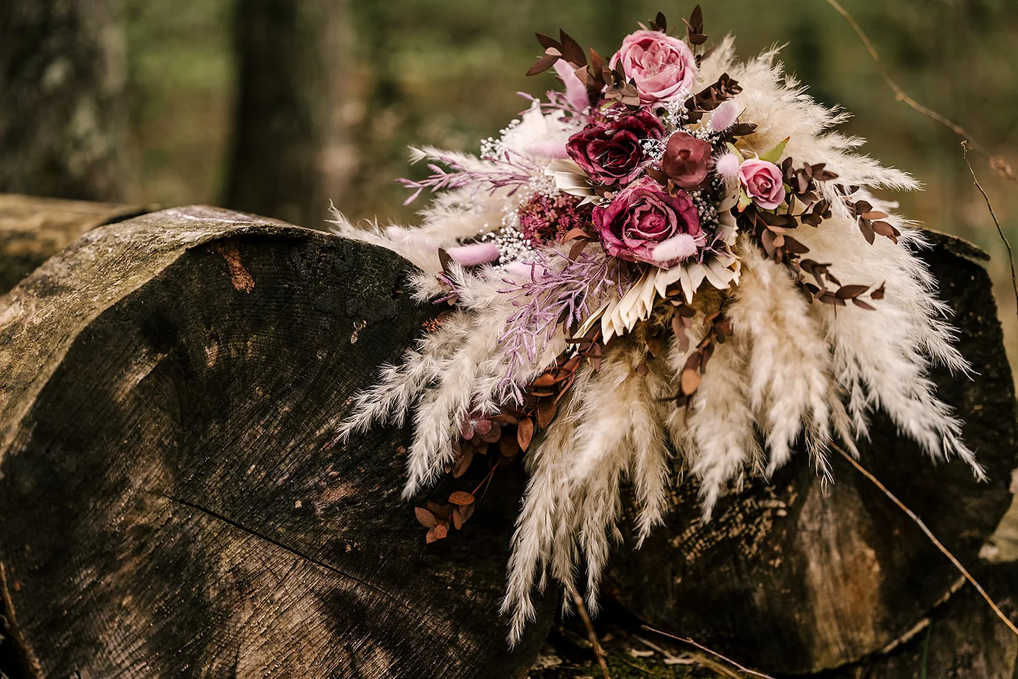 Foxfire Mountain House Wedding DJ: Pink rose and pampas bouquet resting on dark cut logs in a rustic woodland setting.
