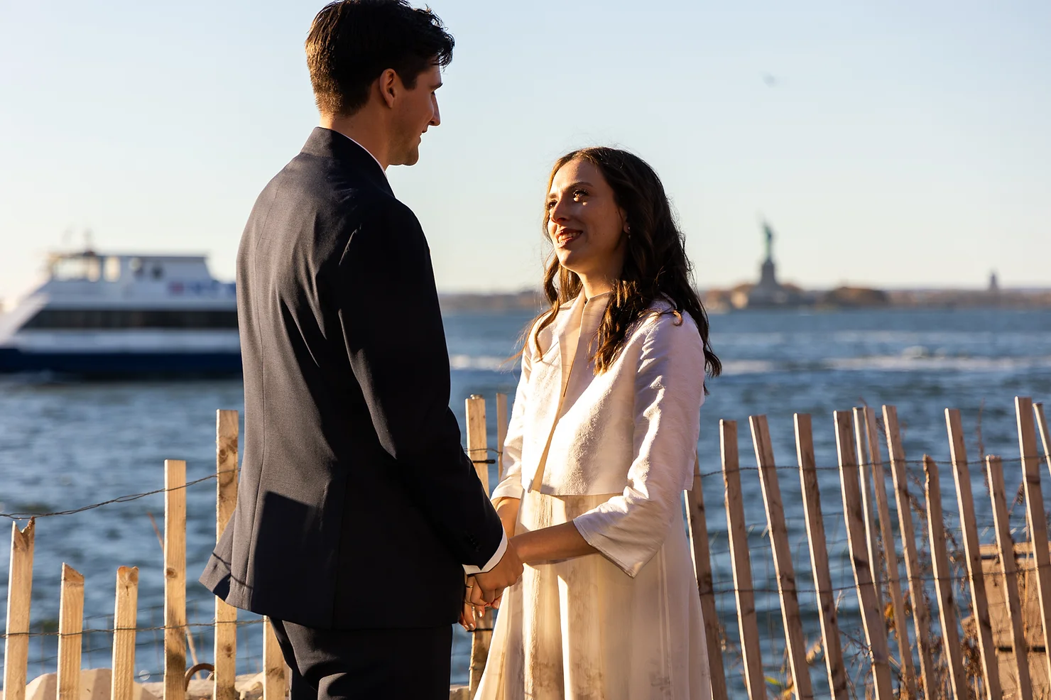 NYC wedding DJ Strong Rope Brewery Couple holding hands by the waterfront at sunset, smiling at each other; ferry and distant Statue of Liberty in the background.