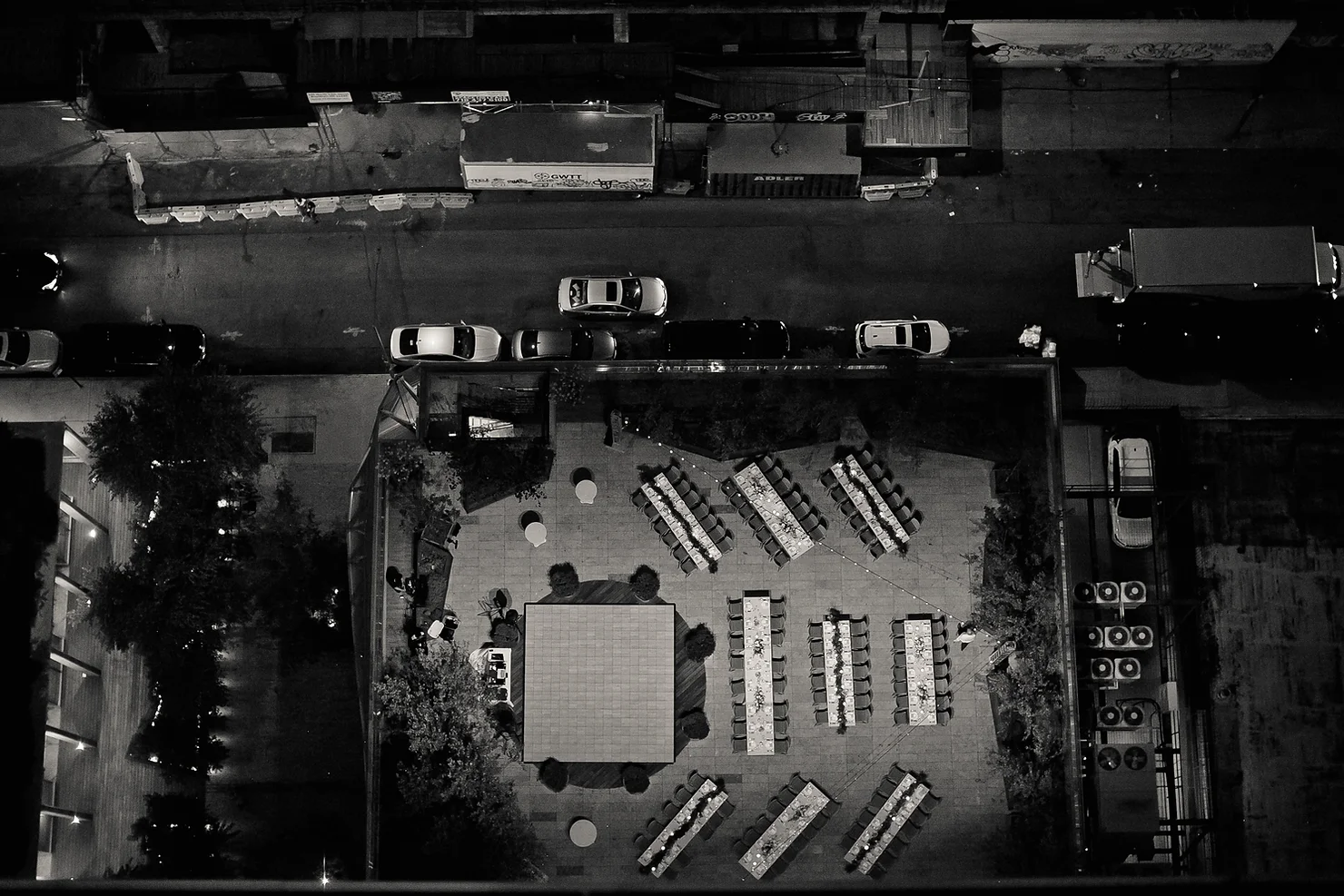 nyc wedding dj the william vale Aerial night view of a black-and-white rooftop dining setup beside a street with parked cars and trucks, quiet urban mood.