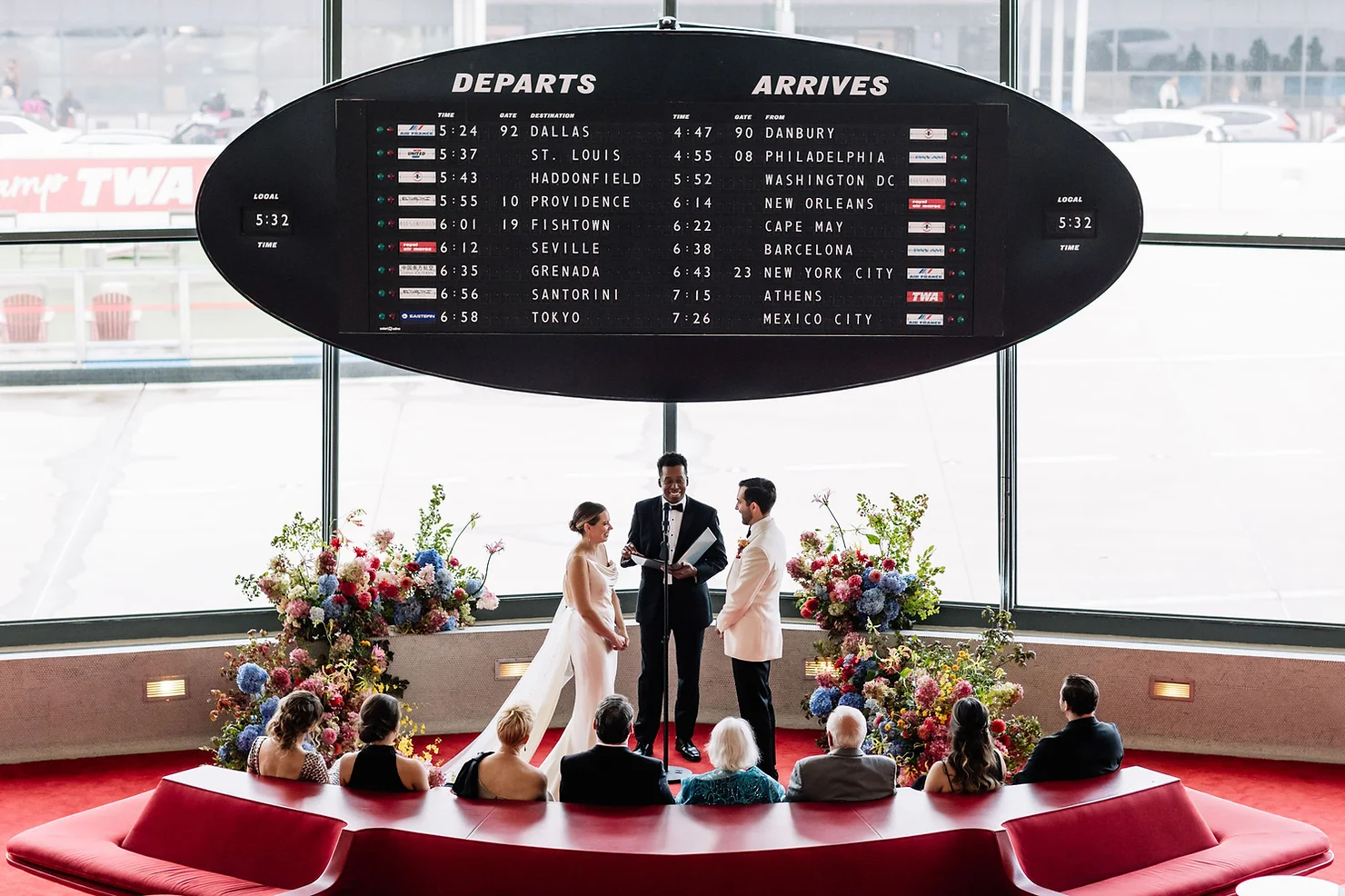nyc non traditional wedding dj Wedding ceremony under a giant DEPARTS/ARRIVES board, with couple and officiant on a red carpet amid colorful flowers.