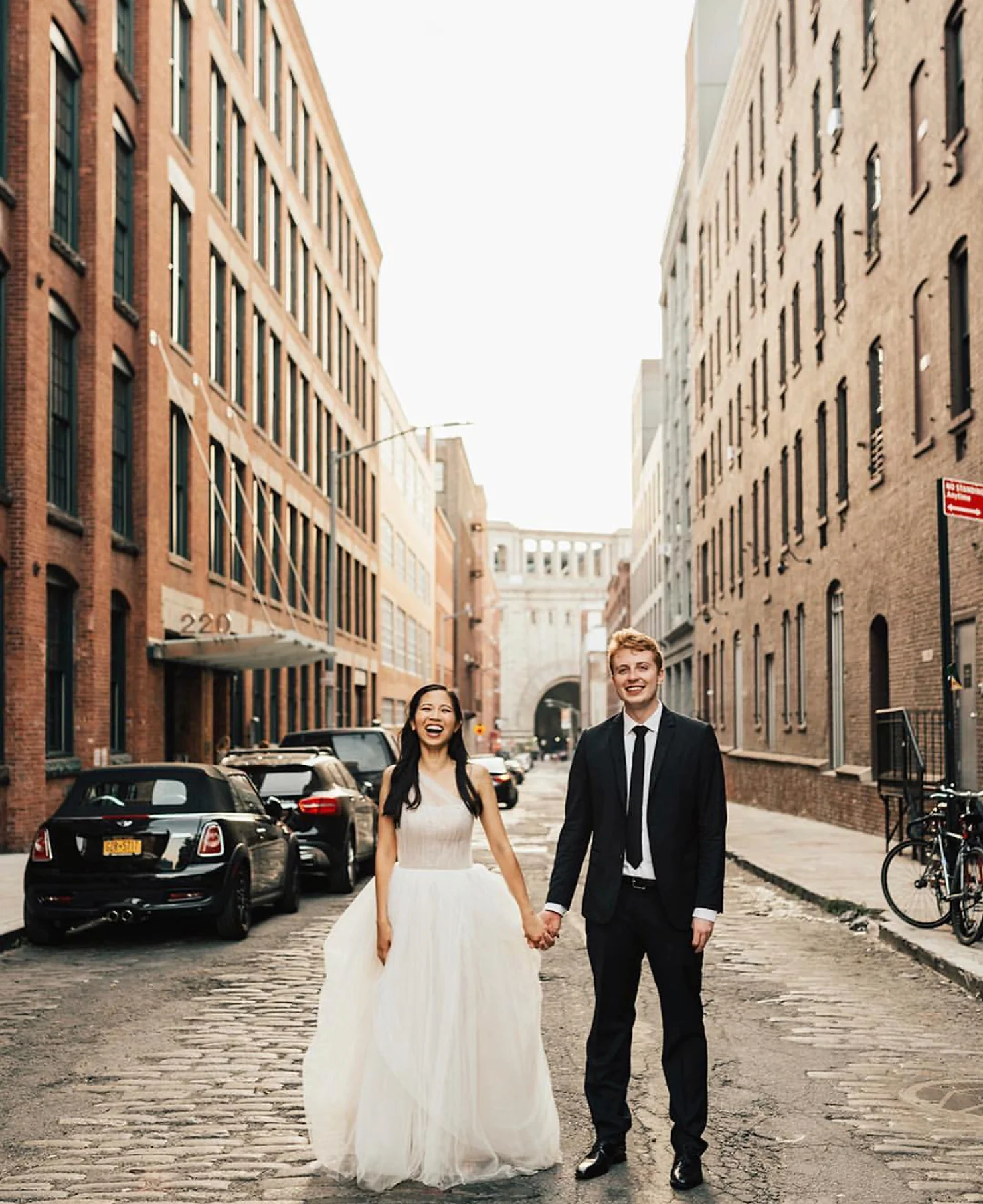 NYC 26 Bridge Wedding DJ Smiling bride in white gown and groom in black suit hold hands on a cobblestone city street between brick buildings.