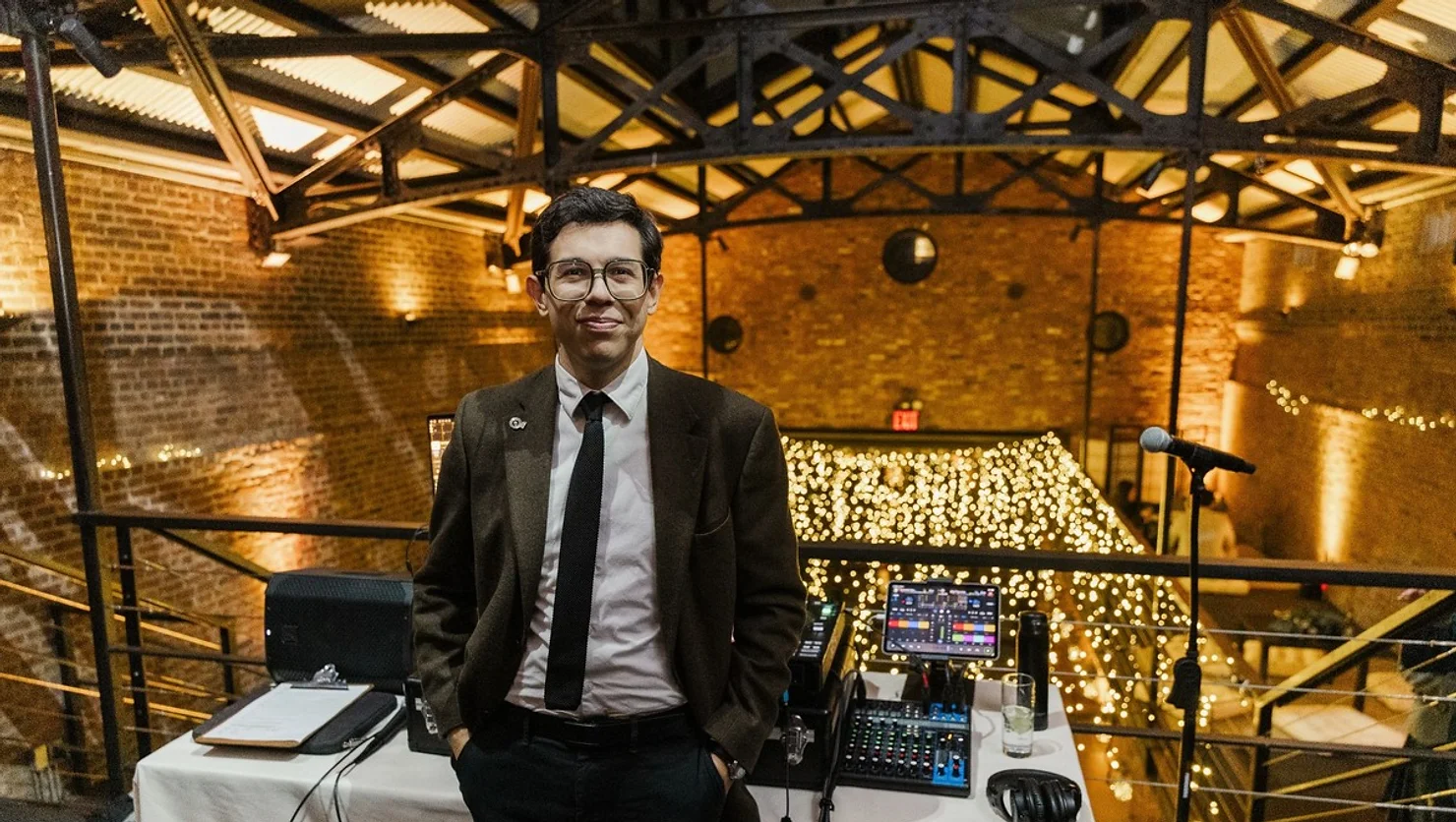 NYC Wedding DJ. Man in a suit stands confidently next to sound equipment. Background features exposed brick, beams, and string lights. Warm, festive ambiance.